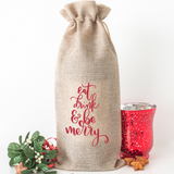 A natural burlap drawstring gift bag with red script text reading 'eat drink & be merry' stands on a white background, styled with a small green holly sprig, red ribbon, and a red candle holder.