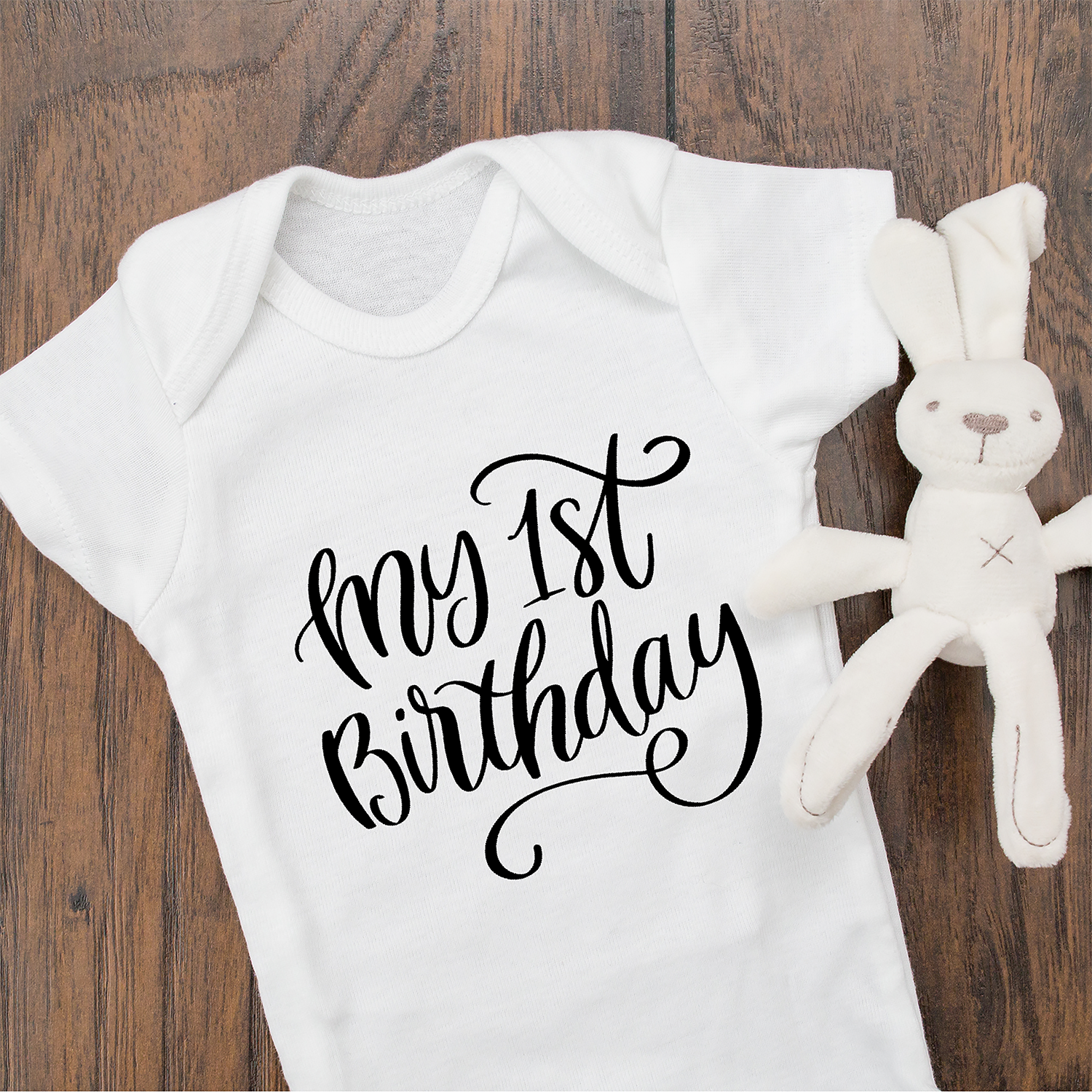 White baby bodysuit with black writing "My 1st Birthday" in script in center of onesie.  Stuffed rabbit toy in white on the right side.