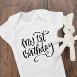 White baby bodysuit with black writing "My 1st Birthday" in script in center of onesie.  Stuffed rabbit toy in white on the right side.