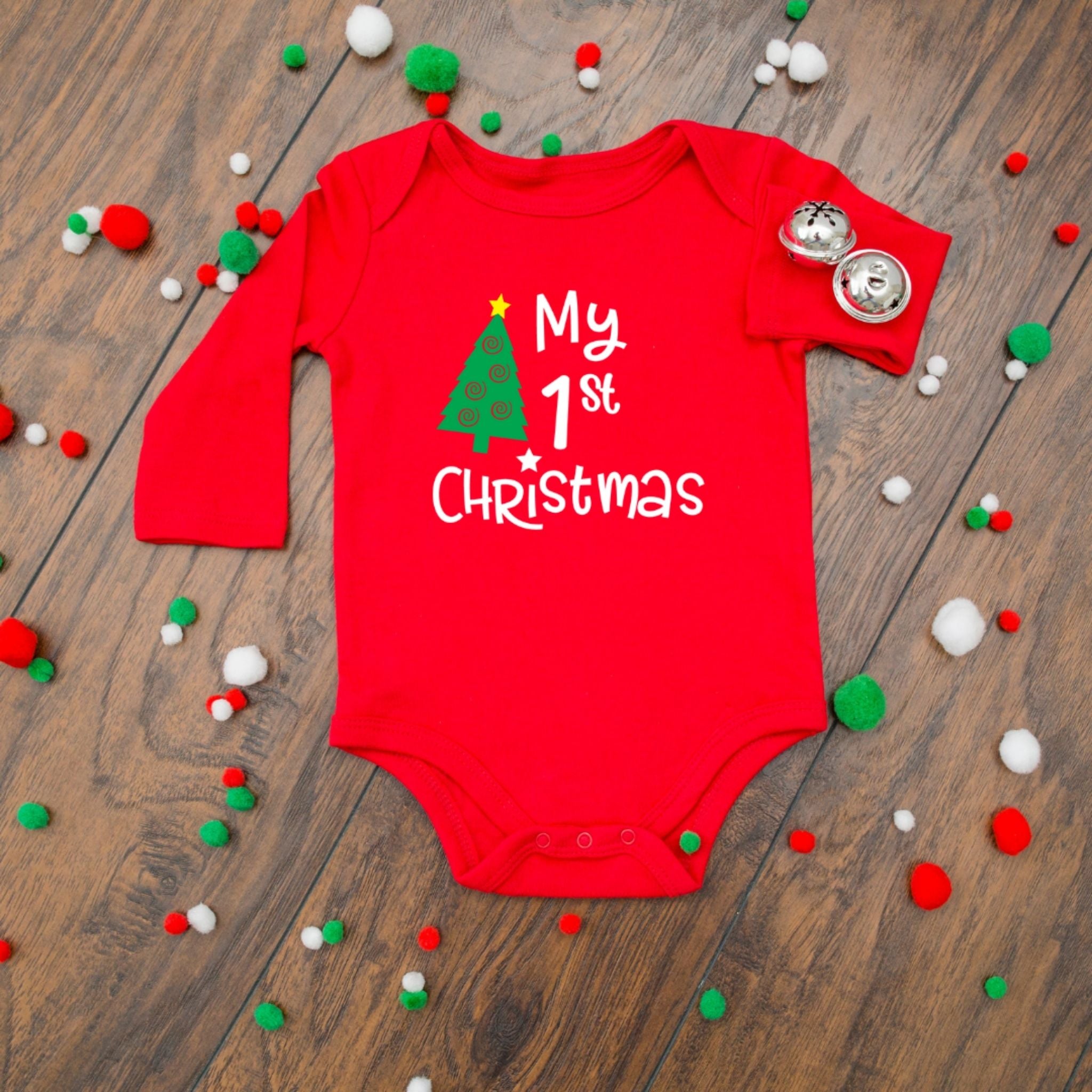 A red long-sleeve baby bodysuit with 'My 1st Christmas' and a green Christmas tree graphic printed on the front, styled flat on a wooden surface with red, green, and white pom-poms and two silver bells.
