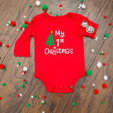 A red long-sleeve baby bodysuit with 'My 1st Christmas' and a green Christmas tree graphic printed on the front, styled flat on a wooden surface with red, green, and white pom-poms and two silver bells.