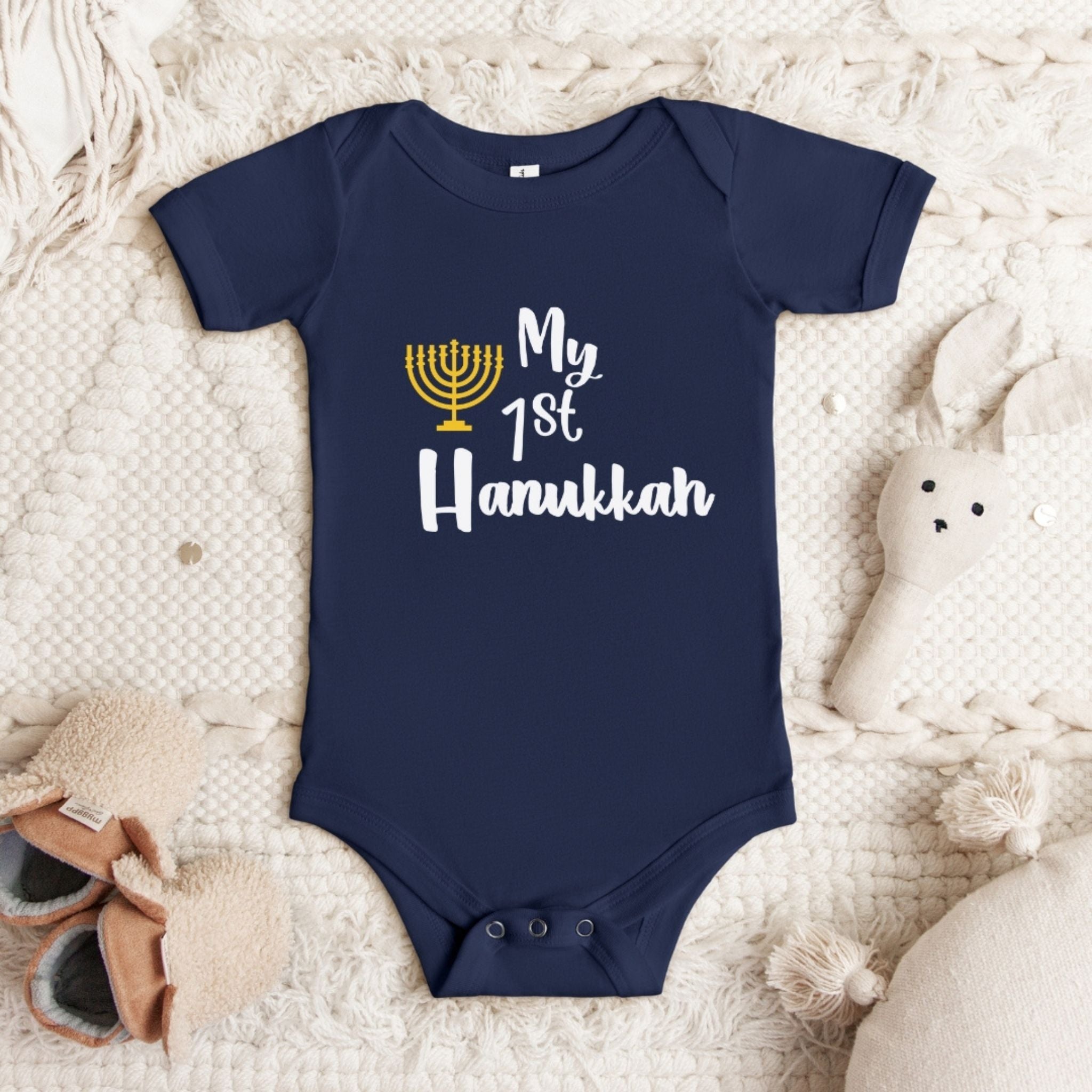 A navy blue short-sleeve baby bodysuit laid flat on a textured cream blanket, with a printed graphic reading 'My 1st Hanukkah' in white and a gold menorah icon. Soft baby props, including a stuffed bunny and small shoes, are placed around it.