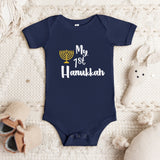 A navy blue short-sleeve baby bodysuit laid flat on a textured cream blanket, with a printed graphic reading 'My 1st Hanukkah' in white and a gold menorah icon. Soft baby props, including a stuffed bunny and small shoes, are placed around it.