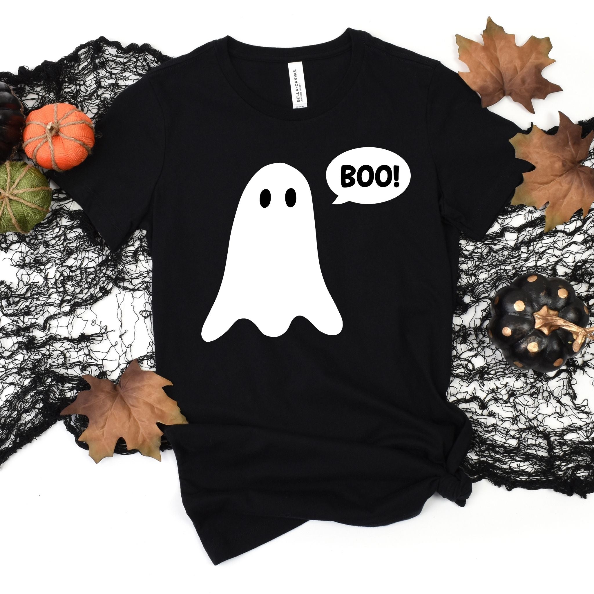 A black short-sleeve t-shirt with a white ghost graphic and a speech bubble that says 'BOO!' on the front, styled in a flat lay with autumn leaves, a small pumpkin, and black lace on a white background.