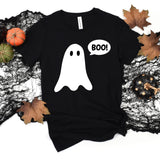 A black short-sleeve t-shirt with a white ghost graphic and a speech bubble that says 'BOO!' on the front, styled in a flat lay with autumn leaves, a small pumpkin, and black lace on a white background.