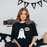 A woman wearing a black t-shirt with a white ghost graphic and a speech bubble that says 'BOO!'. She is sitting on a couch in a cozy, Halloween-themed room with a black cat, a pumpkin, and ghost bunting in the background.