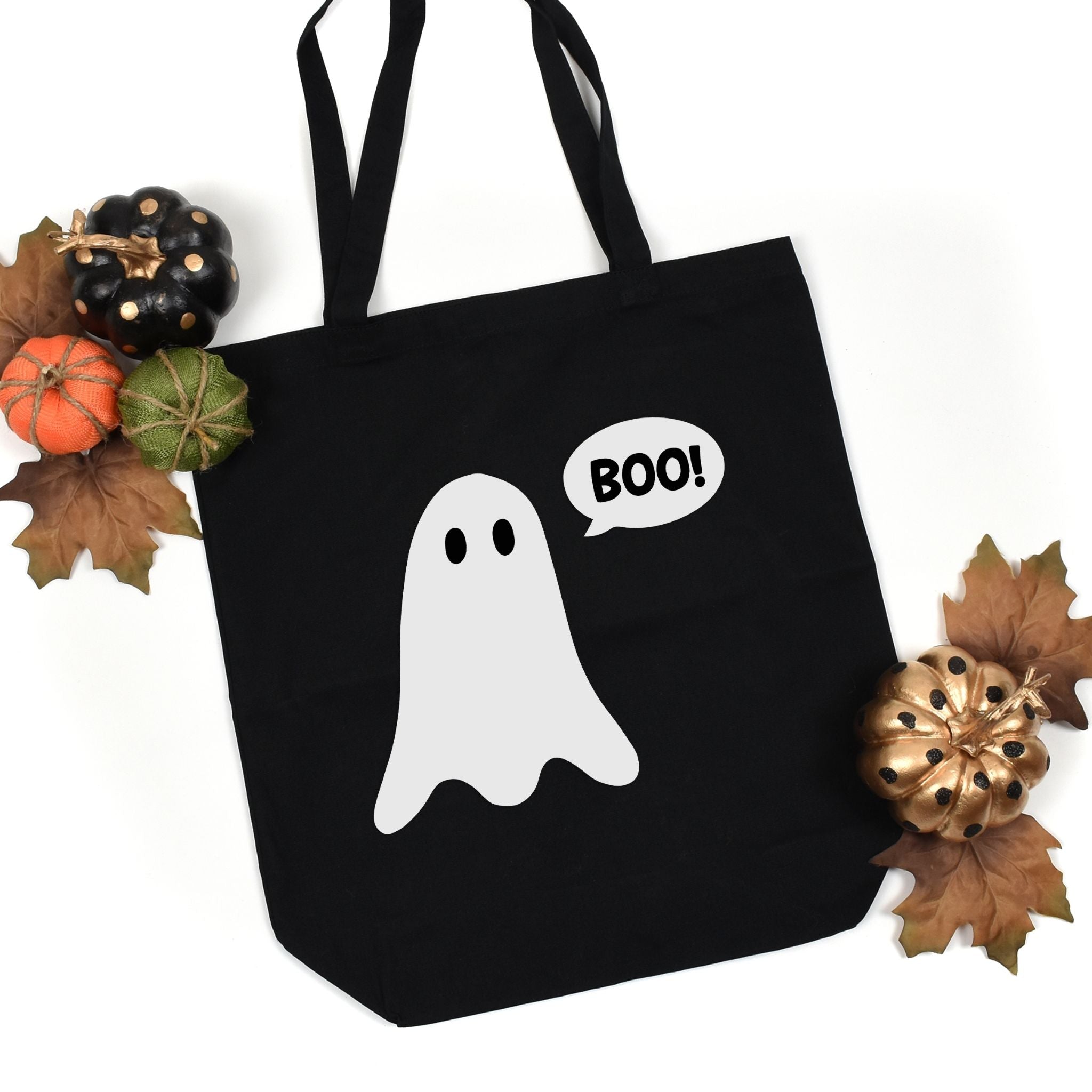 Black canvas tote bag with a white ghost graphic and a speech bubble that says 'BOO!' in white, styled with autumn leaves, small pumpkins, and a gold pinecone on a white background.