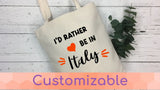 Tote bag with 'I'd rather be in Italy' text on a white wooden surface with greenery.