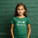 A young girl with long brown hair stands against a dark green brick wall wearing a green t-shirt with 'Irish Blessing' and a shamrock graphic printed in white.