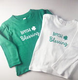 Two folded long-sleeve baby shirts on a light gray background, one green with white 'IRISH Blessing' text and shamrock, and one white with teal 'IRISH Blessing' text and shamrock.