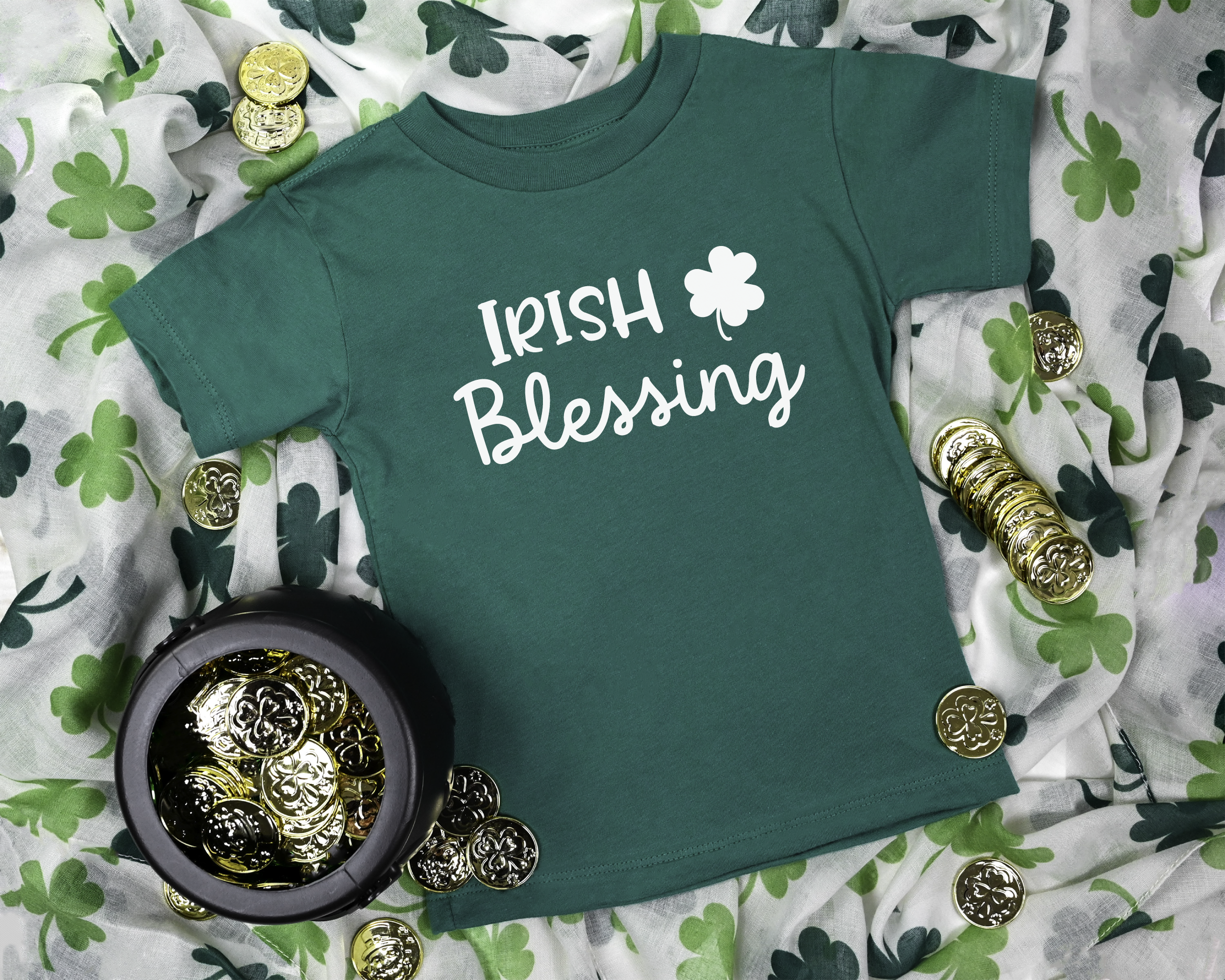 A dark green toddler t-shirt with 'IRISH Blessing' printed in white script and a shamrock graphic, styled in a flat lay with St. Patrick's Day themed props including shamrock-patterned fabric, gold coins, and a small pot of coins.