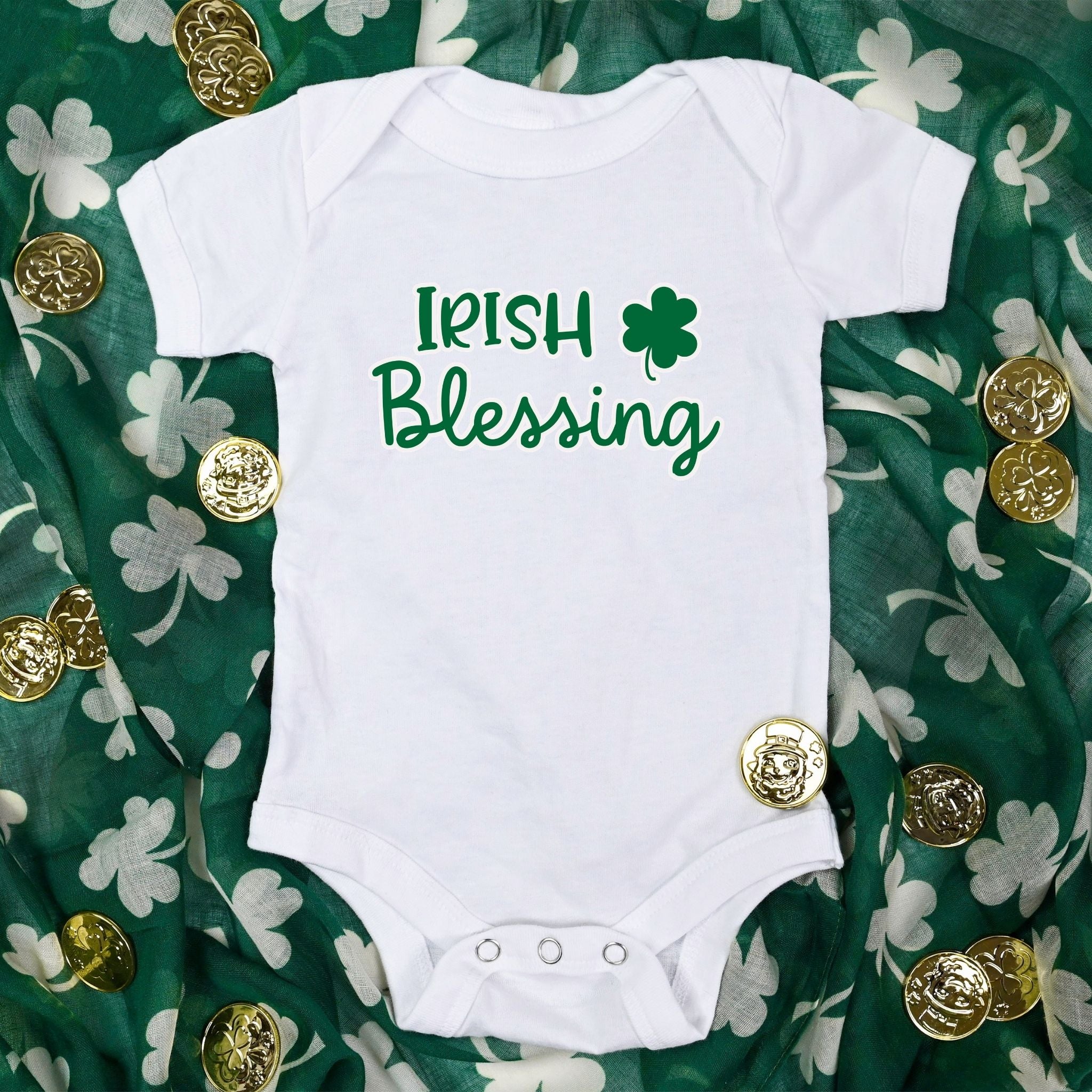 White Baby Bodysuit lying on green scarf with white shamrocks on it and gold coins around the onesie.  Wording on onesie is Irish Blessing with a shamrock in green.