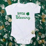 White Baby Bodysuit lying on green scarf with white shamrocks on it and gold coins around the onesie.  Wording on onesie is Irish Blessing with a shamrock in green.
