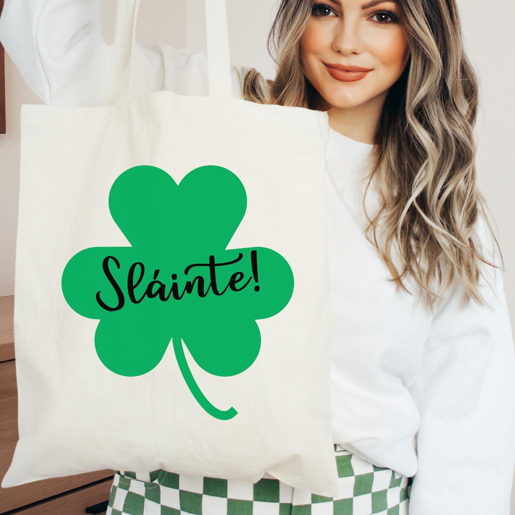A woman holding a cream canvas tote bag with a green shamrock graphic and the word 'Slainte!' in black script, wearing a white sweatshirt and green-and-white checkered pants.