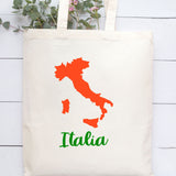 A natural canvas tote bag with a printed orange map of Italy and the word 'Italia' in green script, displayed against a white wood plank background with eucalyptus stems at the top.