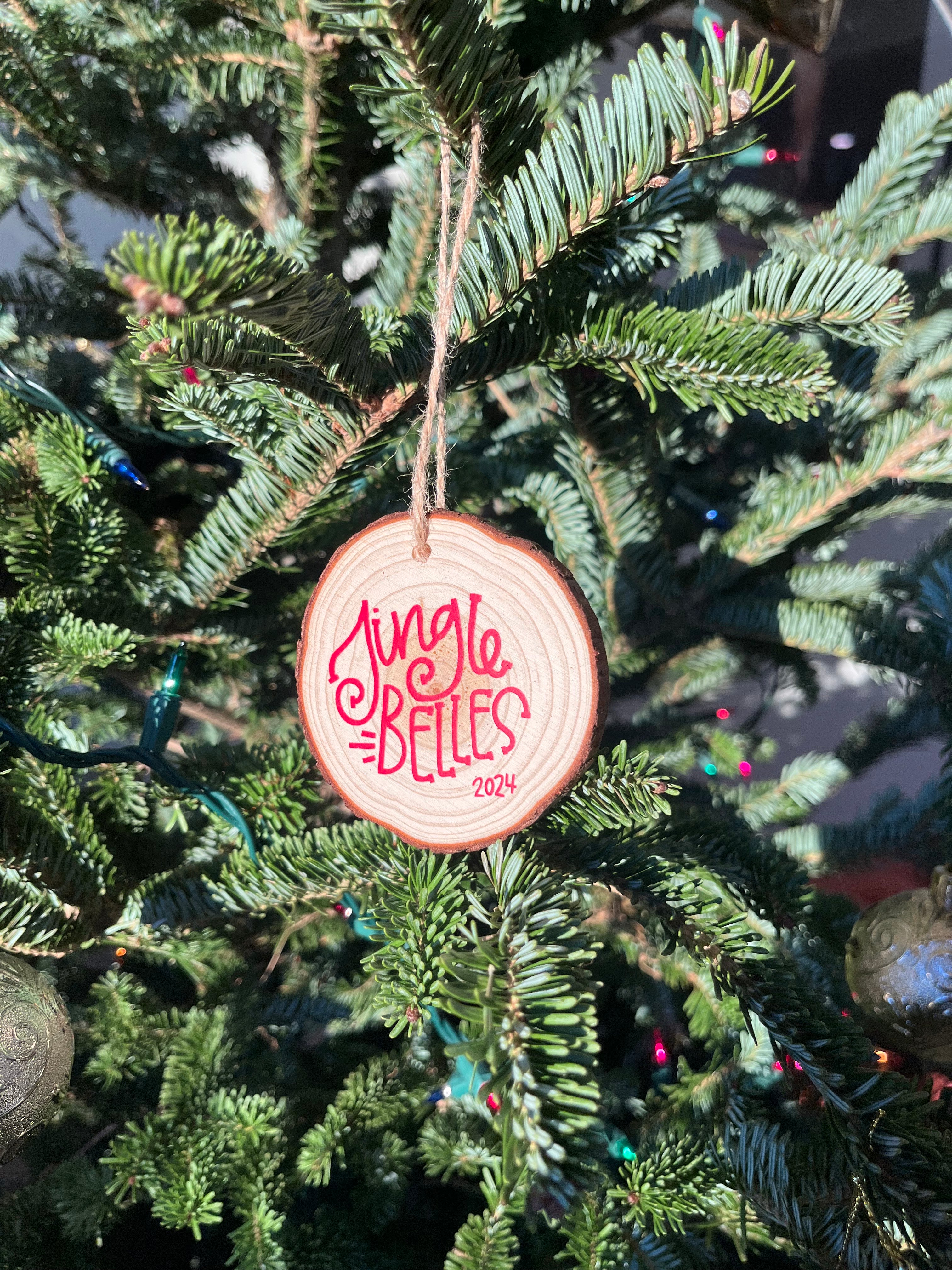 A round wooden Christmas ornament hanging on a green evergreen tree. The ornament is light natural wood with red lettering that reads 'Jingle Bells 2024' and is suspended by a thin string.