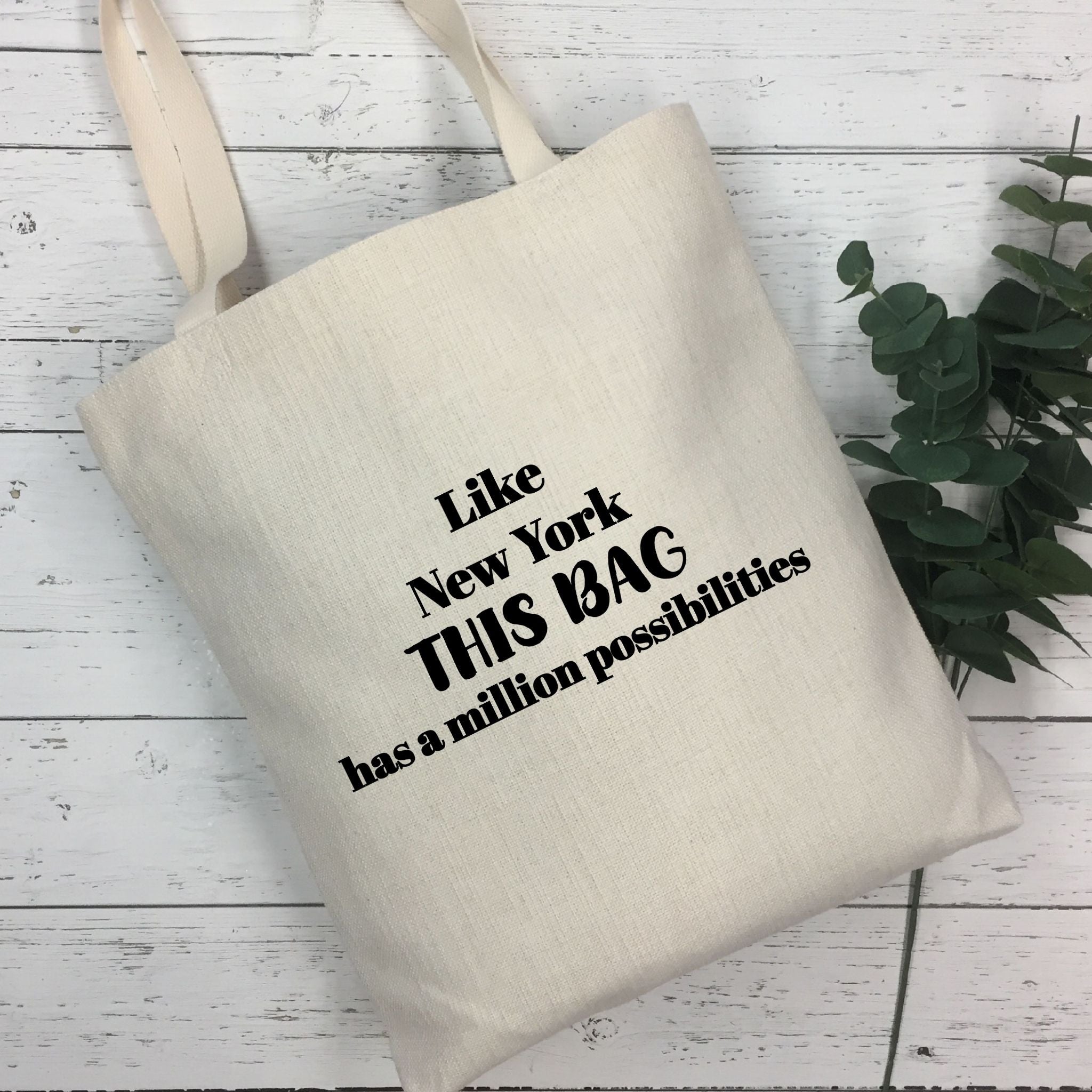 A natural canvas tote bag with black printed text reading 'Like New York THIS BAG has a million possibilities' laid on a white wood plank surface next to green eucalyptus leaves.