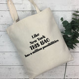 A natural canvas tote bag with black printed text reading 'Like New York THIS BAG has a million possibilities' laid on a white wood plank surface next to green eucalyptus leaves.