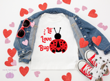 A flat-lay of a white graphic T-shirt with a red ladybug and the words 'I'll Love Bug' in red, styled with an open blue denim jacket, red sunglasses, red and white sneakers, and scattered red and pink glitter hearts on a white background.