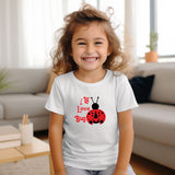 A young girl with curly brown hair smiling in a white t-shirt featuring a red ladybug graphic and the text 'I love bugs'. The photo is taken in a brightly lit living room with a sofa in the background.