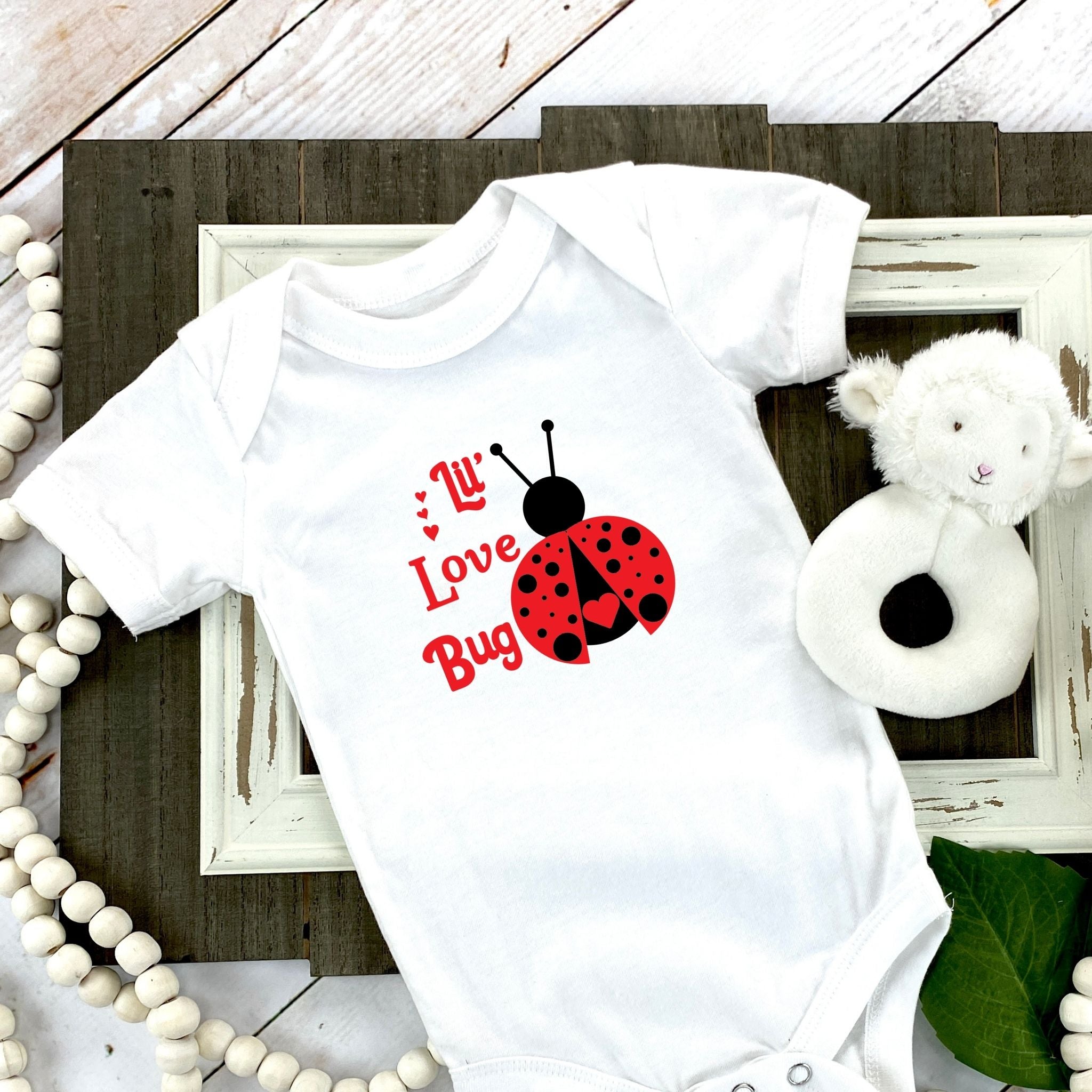 White baby onesie laid down on wood frame with sheep rattle on the right with red working stating Lil' Love Bug and a red and black ladybug on the right. 