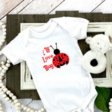 White baby onesie laid down on wood frame with sheep rattle on the right with red working stating Lil' Love Bug and a red and black ladybug on the right. 