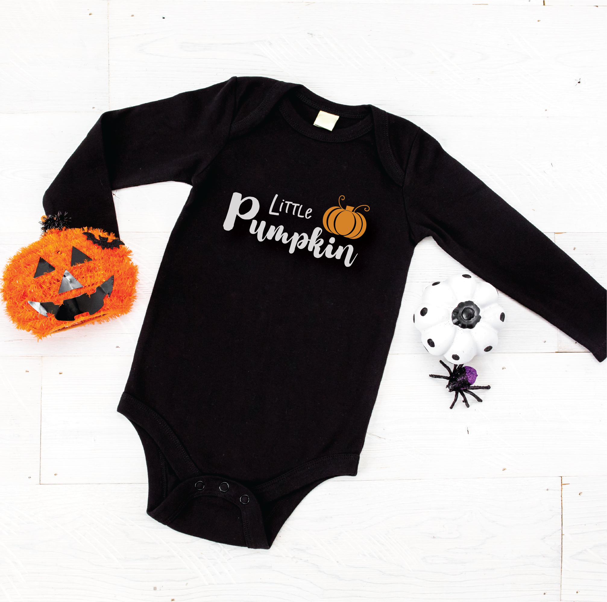 A black long-sleeve baby bodysuit with 'Little Pumpkin' printed in white script and a small orange pumpkin graphic, laid flat on a white wood background with Halloween props.