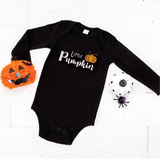 A black long-sleeve baby bodysuit with 'Little Pumpkin' printed in white script and a small orange pumpkin graphic, laid flat on a white wood background with Halloween props.
