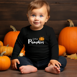 A baby sitting on a wooden floor surrounded by pumpkins, wearing a black long-sleeve shirt with 'Little Pumpkin' printed in white script and a small orange pumpkin graphic.