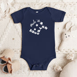 A navy blue short-sleeve baby bodysuit with white text reading 'Made in BROOKLYN' and stars, laid flat on a cream knit blanket with baby shoes and a plush toy nearby.