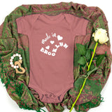A dusty rose baby bodysuit with 'Made in Brooklyn' printed in white on the chest, styled flat-lay on a green patterned shawl with a white rose and wooden toy.