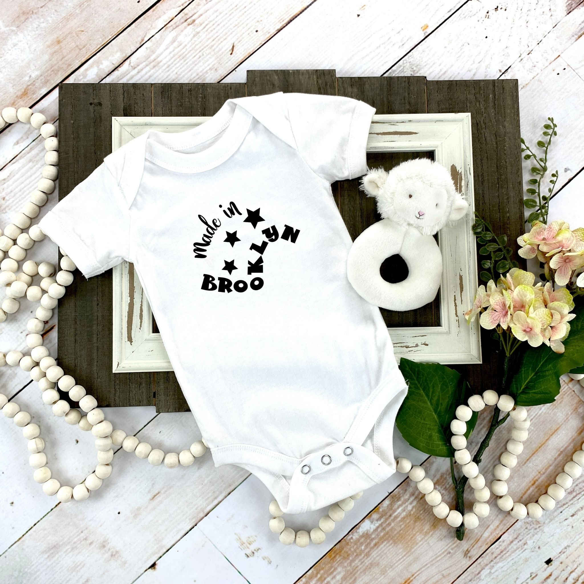 A white short-sleeve baby bodysuit with a black 'Made in Brooklyn' graphic, styled in a flat-lay arrangement on a rustic white wood background with a teddy bear, flowers, and bead garlands.