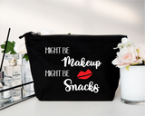 A black canvas cosmetic pouch with a zipper closure, featuring the text 'MIGHT BE Makeup MIGHT BE Snacks' in white script with a red lips graphic. The pouch is styled on a white surface with decorative props, including a Chanel perfume bottle, a glass reed diffuser, and white and pink flowers.