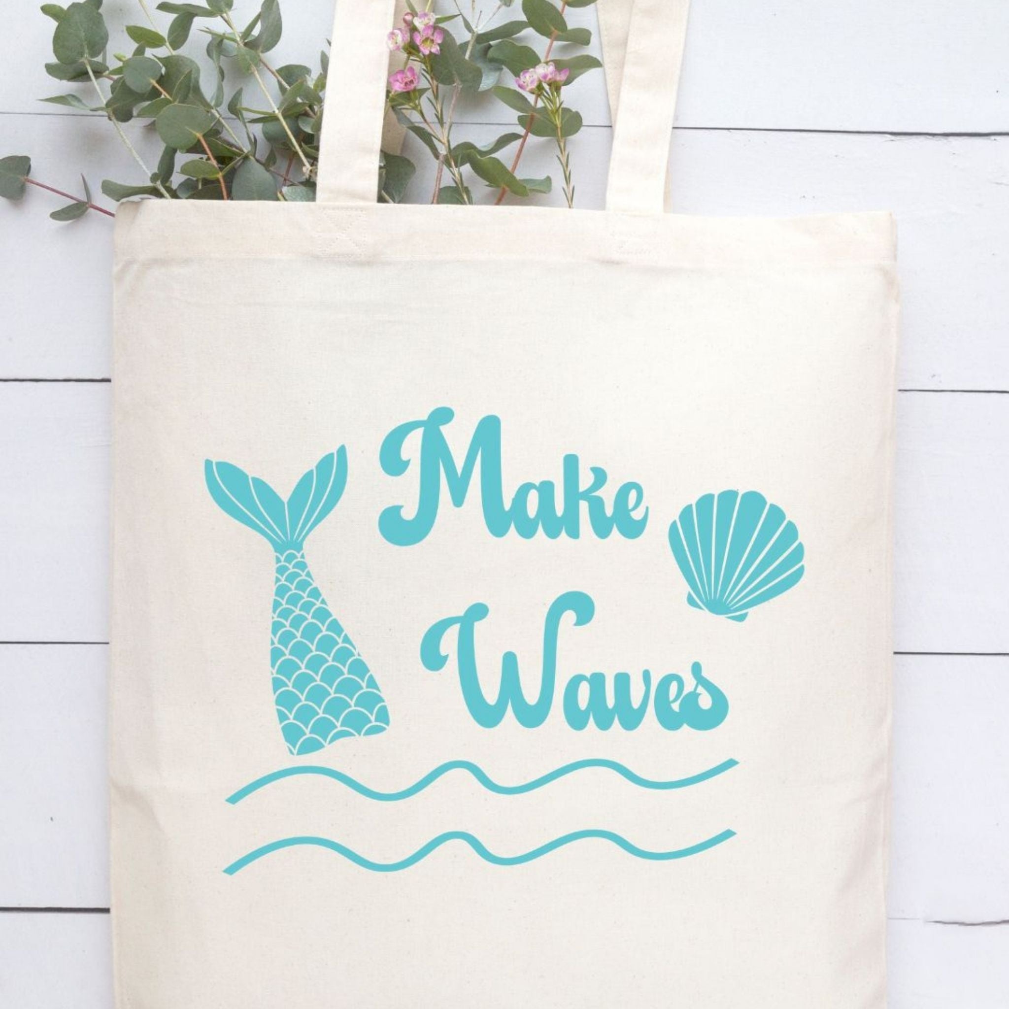 An off-white canvas tote bag with a teal mermaid-themed graphic that reads 'Make Waves' alongside a mermaid tail fin, seashell, and wavy lines, displayed against a white wood plank background with green leaves at the top.