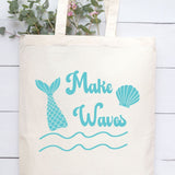 An off-white canvas tote bag with a teal mermaid-themed graphic that reads 'Make Waves' alongside a mermaid tail fin, seashell, and wavy lines, displayed against a white wood plank background with green leaves at the top.