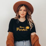 A woman wearing a black graphic t-shirt with 'Mama Pumpkin' in yellow text and a pumpkin illustration, styled with a brown wide-brim hat, a brown cardigan, and light blue jeans against a light gray background.