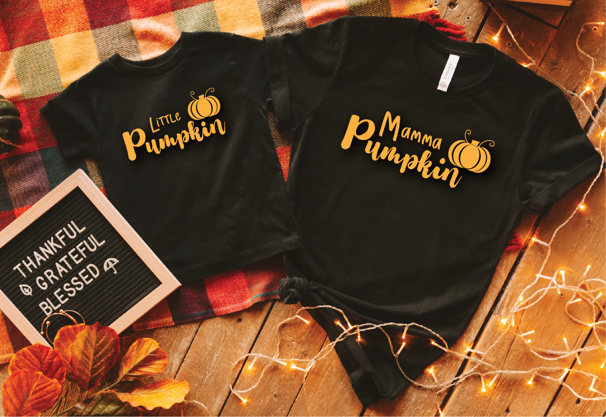 Two black short-sleeve T-shirts laid flat on a wooden surface with a plaid blanket, fall leaves, and string lights. The smaller shirt reads "Little Pumpkin" and the larger shirt reads "Mamma Pumpkin," both with small pumpkin graphics.