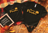 Two black short-sleeve T-shirts laid flat on a wooden surface with a plaid blanket, fall leaves, and string lights. The smaller shirt reads "Little Pumpkin" and the larger shirt reads "Mamma Pumpkin," both with small pumpkin graphics.
