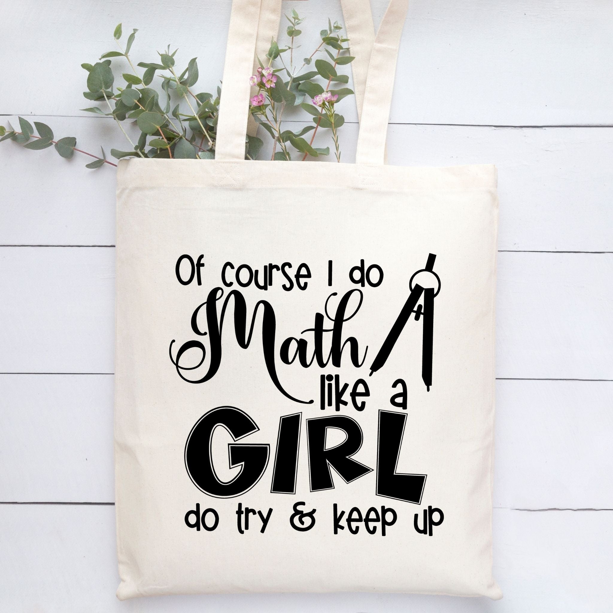 An off-white canvas tote bag with black printed text that reads 'Of course I do Math like a GIRL do try & keep up' and a chalkboard graphic, hanging against a white wood plank background with green leaves at the top.
