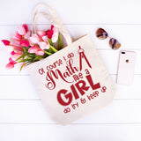A beige canvas tote bag with red text reading 'Of course I do Math like a GIRL do try & keep up' and a red compass graphic, styled on a white wood background with pink tulips, sunglasses, and a smartphone.