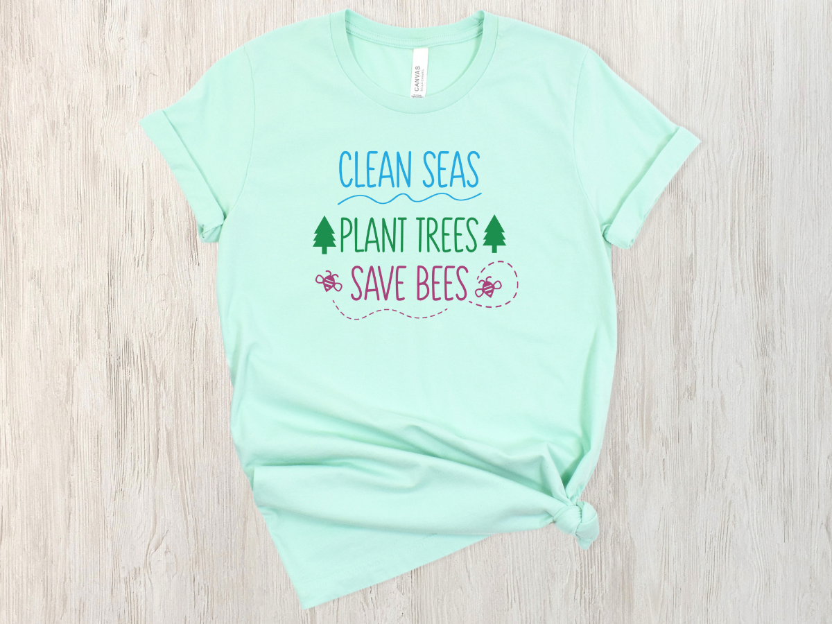 A mint green short-sleeve t-shirt with a colorful graphic print reading 'CLEAN SEAS PLANT TREES SAVE BEES' in blue, green, and pink, styled flat on a light wood background.