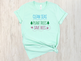 A mint green short-sleeve t-shirt with a colorful graphic print reading 'CLEAN SEAS PLANT TREES SAVE BEES' in blue, green, and pink, styled flat on a light wood background.