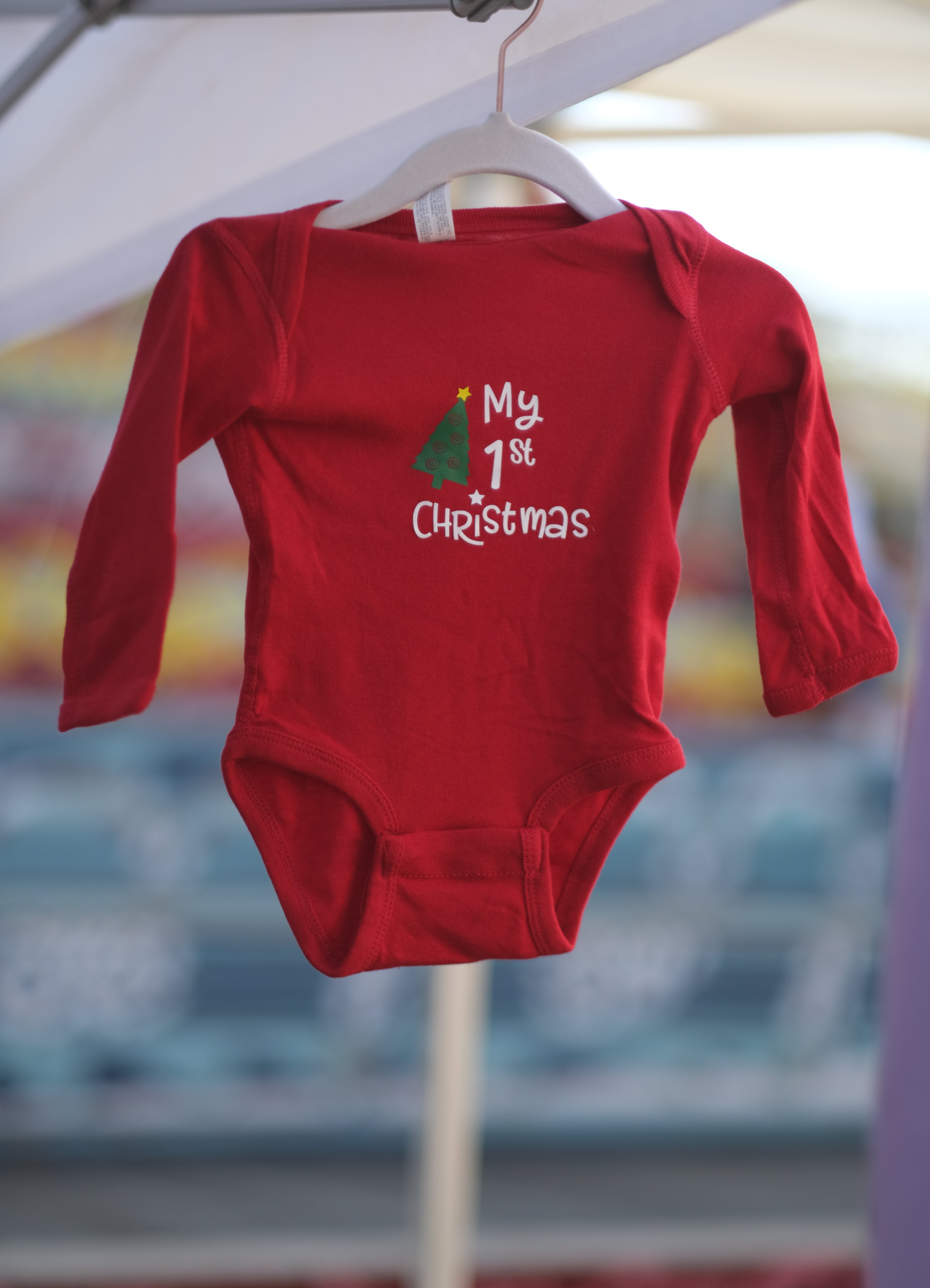 Red long sleeve baby bodysuit  hanging from hanger with white "my 1st Christmas" writing and green tree tilted to the right with a red star topper.