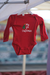 Red long sleeve baby bodysuit  hanging from hanger with white "my 1st Christmas" writing and green tree tilted to the right with a red star topper.