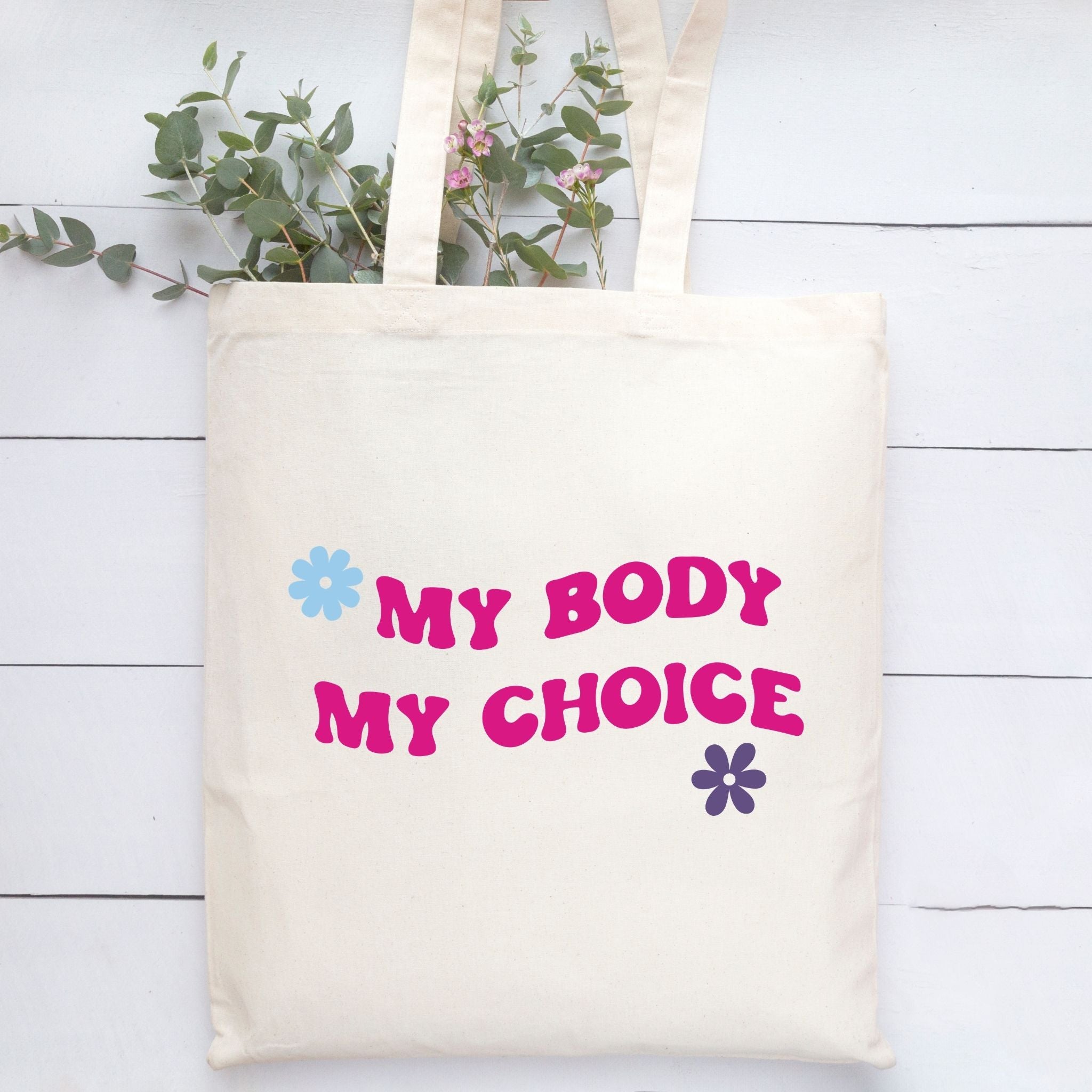 An off-white canvas tote bag with the text 'MY BODY MY CHOICE' printed in pink bubble letters, accented with a light blue flower on the left and a dark purple flower on the right. The bag is displayed on a white wooden plank surface with green eucalyptus stems peeking out from the top.