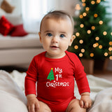 A baby wearing a red long-sleeve bodysuit with a 'My 1st Christmas' graphic, sitting in a cozy living room with a blurred Christmas tree in the background.
