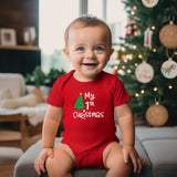 A baby wearing a red short-sleeve bodysuit with 'My 1st Christmas' and a green tree graphic, sitting on a gray sofa in front of a decorated Christmas tree.