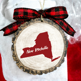 A round wood slice ornament with a rustic bark edge, featuring a red silhouette of New Rochelle and the words 'New Rochelle' in white script. The ornament is tied with a red and black buffalo plaid ribbon bow and hangs against a white fabric background with a hint of red at the bottom.