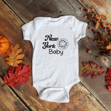 A white short-sleeve baby bodysuit laid flat on a rustic wooden surface, printed with the words 'New York Baby' and a donut graphic in black. The scene is styled with autumn leaves, a small pumpkin, and dried berry sprigs around the garment.