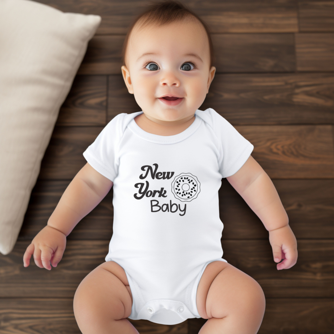 A baby lying on a dark wood floor wearing a white short-sleeve bodysuit with a black 'New York Baby' graphic and donut illustration. A cream pillow is visible on the left side.